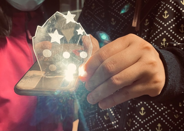 Hände halten eine transparente, mit Sternen und Kreisen verzierte Platte über eine leuchtende Smartphone-Taschenlampe.